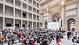 In einem teilweise fertiggestellten Innenraum sieht sich ein Publikum eine Präsentation an