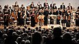 Ein Orchester steht vor einem Publikum auf der Bühne