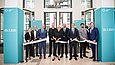 Mehrere Menschen halten ein weißes Band und Scheren in der Hand.