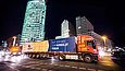 Vor der Kulisse des nächtlichen Berlins fährt ein mit einer großen Kiste beladener LKW eine Straße entlang