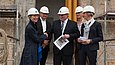 Fünf Menschen mit weißen Bauhelmen stehen auf einer Baustelle, ein Mann in der Mitte schüttelt einer Frau links die Hand