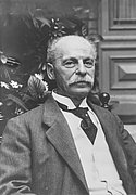 Historisches Schwarz-Weiß-Porträt von Eduard Arnhold. Der Mann mit Schnurrbart sitzt im Anzug vor einer Innenraumkulisse und blickt in die Kamera.