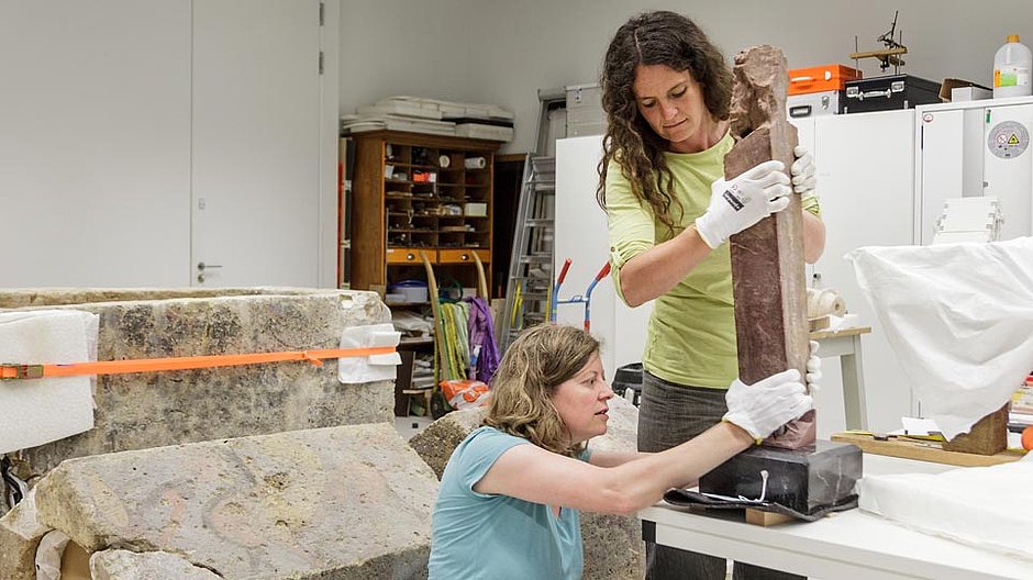 Two women mount an ancient object on a pedestal
