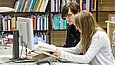 Zwei junge Menschen sitzen an einem Tisch und blicken in ein Buch, vor ihnen ein Computerbildschirm