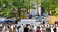 Ein Mann steht auf der Treppe einer Villa im Freien und spricht in ein Mikrofon zu einem Publikum