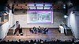 Podiumssituation mit einer parkettdominierten Bühne vor einem besetzten Auditorium