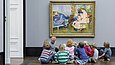 A woman and a group of children sit on the floor in front of a painting