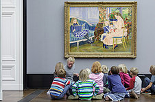 A woman and a group of children sit on the floor in front of a painting