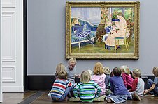 A woman and a group of children sit on the floor in front of a painting