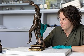 Dorothea Habel reinigt eine Bronzefigur mit einem feinen Stäbchen und trägt dabei grüne Handschuhe.