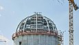 Blick auf eine sich im Bau befindlichen Gebäudekuppel, über der ein grüner Kranz an einem Kran schwebt