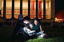 Zwei junge Personen sitzen an Abend vor einem prachtvollen Museum und beleuchten mit Taschenlampen ein Programmheft