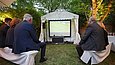 Menschen sitzen auf Bierbänken in einem Garten und schauen auf einer großen Leinwand ein Fußballspiel an