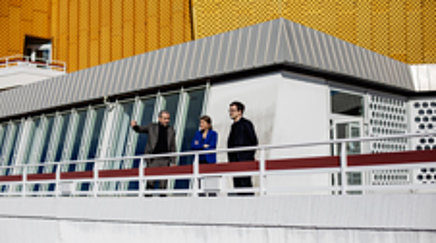 Drei Personen stehen auf der Terrasse eines modernen Gebäudes.