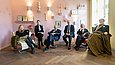 In einem als Wohnraum gestalteten Ausstellungsraum sitzen 6 Personen auf Möbeln