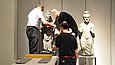 Drei Menschen bewegen eine Skulptur in einem Ausstellungraum