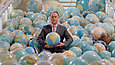 Mann mit Anzug und Krawatte steht in einem Meer von Globen und hält einen Globus hoch