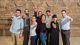Drei Frauen und fünf Männer posieren lächelnd in einem Museum und zeigen mit den Fingern auf die Kamera