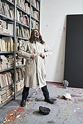 Jonathan Meese steht lachend in einem hellen Mantel in seinem Atelier vor Bücherregalen und einer großformatigen schwarzen Leinwand.