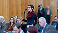 Eine Frau steht zwischen mehreren Menschen in einer Stuhlreihe und spricht in ein Mikrofon