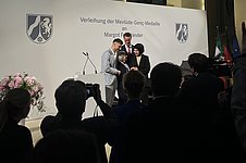 Zwei Männer und eine Frau beglückwünschen eine ältere Dame auf einer Bühne