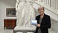 Eine Frau steht neben der Skulptur zweier Frauen in einem Treppenhaus und hält eine kleine blaue Pappschachtel in den Händen