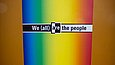 Regenbogenfarbiges Plakat mit der englischen Aufschrift "We (all) are the people"