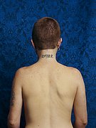 A rear view of a naked person with closely cropped hair and the tattoo ‘DYKE’ on the nape of their neck, set against a blue patterned background.