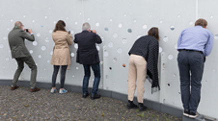 Mehrere Menschen blicken und fotografieren durch eine Wand mit Löchern.