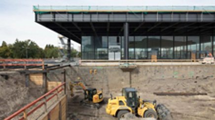  Excavator in a pit in front of a modern building.