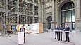 Eine Frau links im Bild spricht an einem Rednerpult, rechts stehen drei Männer an einem langen Tisch. Im Hintergrund sieht man die eingerüstete Fassade eines Gebäudes