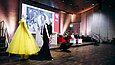 Bunte Kleider vor einer Bühne mit großer Leinwand und Menschen auf Sofa im Hintergrund