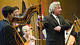 A conductor standing among musicians next to a harp