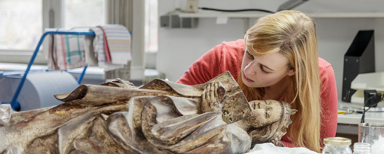 Eine junge Frau beugt sich über eine Heiligenskulptur aus Lindenholz