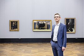 Ein Mann im Anzug steht in einem Ausstellungssaal vor drei Gemälden des 19. Jahrhunderts