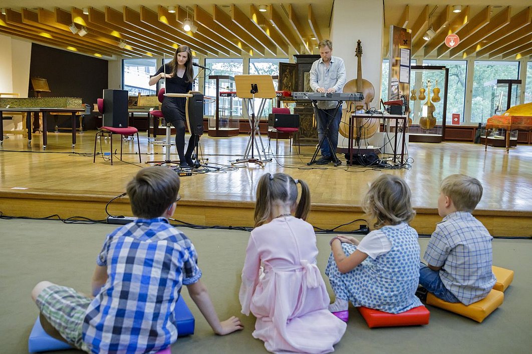 Kinder bei einem Konzert im Musikinstrumenten-Museum. Foto: SPK/photothek.net/Thomas Trutschel Vier Kinder sitzen vor einer Bühne, auf der eine Frau Theremin spielt und ein Mann Keyboard.