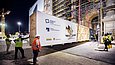 Mehrere Arbeiter mit Bauhelmen beobachten, wie bei Nacht eine große Holzkiste durch die Pforte eines eingerüsteten Gebäudes gebracht wird