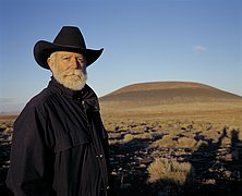 James Turrell mit schwarzem Mantel und breitkrempigem Hut vor der Landschaft des Roden Crater in Arizona.