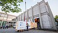 Eine große Holzkiste wird aus einem Gebäude durch ein Rolltor nach draußen geschoben und mit Spanngurten an einem Kran befestigt, während mehrere Personen im Hintergrund das Spektakel beobachten