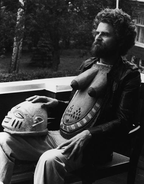 Fotografie von Leonore Mau, auf der ein sitzender Mann in einem leichten Seitenprofil zu sehen ist. Er trägt vor der Brust eine Art Brustpanzer. Eine Maske liegt auf seinem rechten Knie.