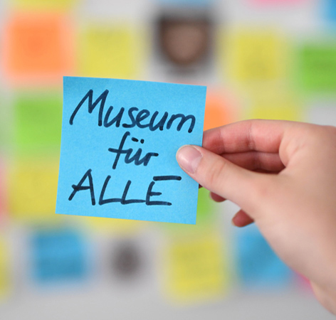 Digitale Partizipation Eine Hand hält ein Post-it mit der Aufschrift "Museum für alle" vor einer Wand mit bunten Post-its hoch