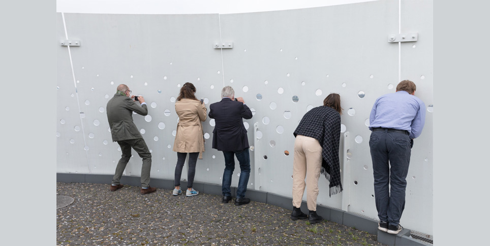Mehrere Menschen blicken und fotografieren durch eine Wand mit kleinen Löchern