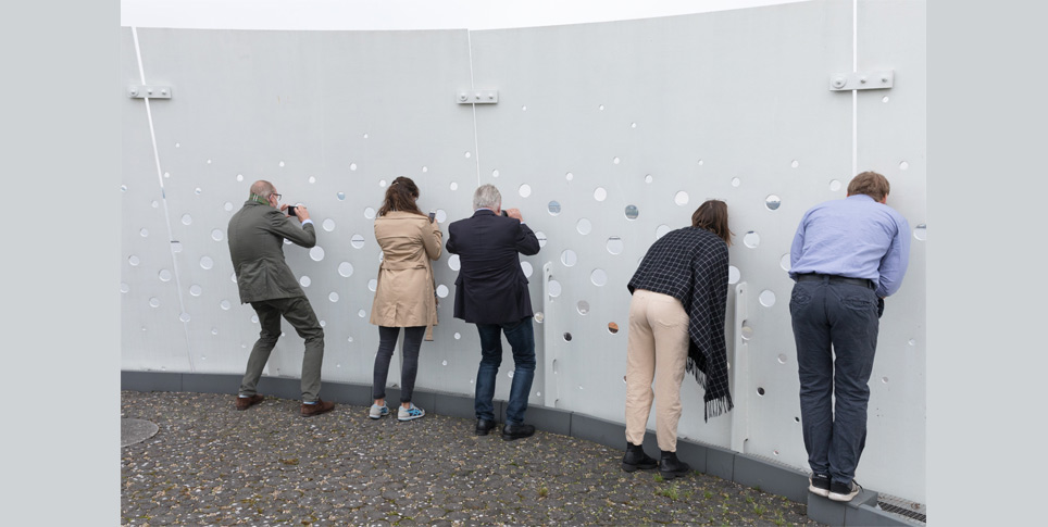 Digitale Partizipation Mehrere Menschen blicken und fotografieren durch eine Wand mit kleinen Löchern