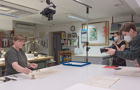 Eine Frau bearbeitet an einem Tisch ein historisches Dokument und wird dabei von zwei Personen fotografiert.