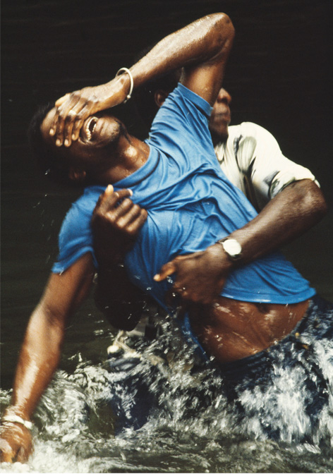Vorbereitungen zum Regenopfer am Grand Etang See, einer Zeremonie des Shangokults, 1979 © bpk / S. Fischer Stiftung / Leonore Mau Zwei Männer in T-Shirt und Jeans stehen bis zur Hüfte im Wasser, der hintere hält den vorderen, sich in Trance windenden an der Hüfteumfasst