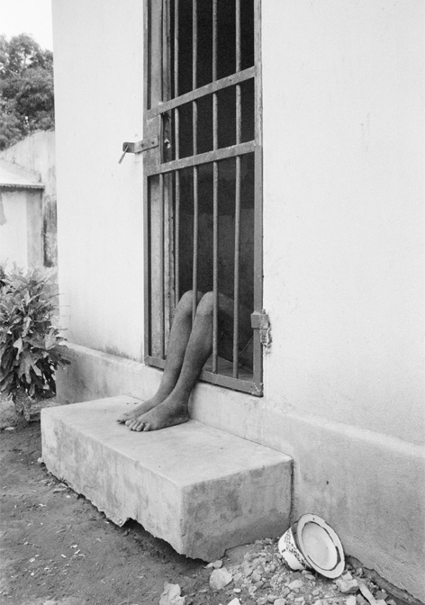 ein Paar nackte Beine ragen bis zu den Knien durch eine Gittertür eines einfachen, weißen Hauses auf Sandboden