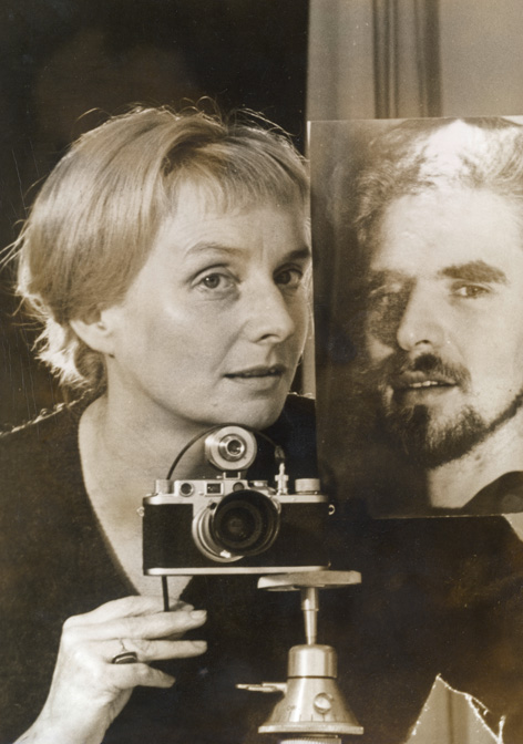 Selbstporträt von Leonore Mau mit einem Porträtfoto von Hubert Fichte, 1962 © bpk / S. Fischer Stiftung / Leonore Mau Sepia-Porträt einer blonden Frau mit älterer Kamera auf Tischstativ neben lebensgroßem Fotoporträt eines vollbärtigen Mannes
