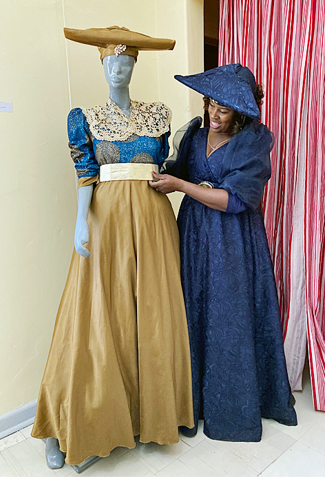 Modesdesignerin Cisla Rukoro mit der von ihr entworfener Herero-Tracht Eine Frau mit langem Kleid und horizontal sehr ausladendem Hut steht stolz neben einer Modepuppe in ähnlicher Bekleidung