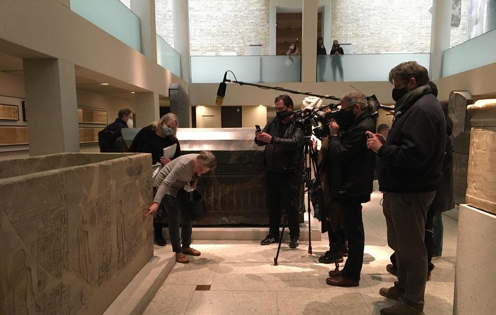 Museumsdirektorin Friederike Seyfried zeigt der Presse im Neuen Museum die Flecken an verschiedenen Objekten Eine Gruppe von Journalisten steht um eine Frau, die auf eine Stelle auf einem Steinsarkophag zeigt
