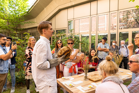 Foodperfomance von Chmara.rosinke im Garten des Kunstgewerbemuseums © Stefanie Heider Menschen gruppieren sich um einen gedeckten Holztisch in einem begrünten Innenhof um einen Mann, der ein Brot in der Hand hält